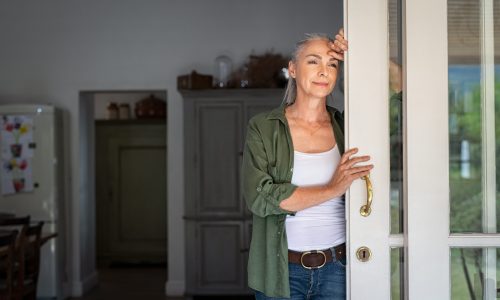 Thoughtful senior woman standing at home entrance leaning against door. Mature woman waiting husband at doorway and looking away. Lady in casual clothing standing at door and thinking.