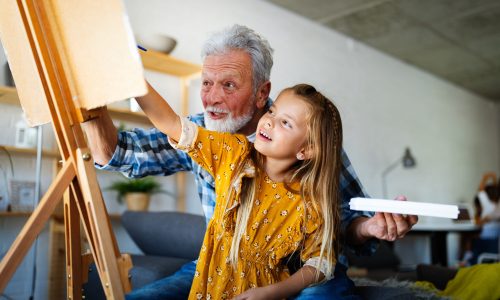 Grandfather spending happy time with granddaughter. Senior man with child painting on canvas