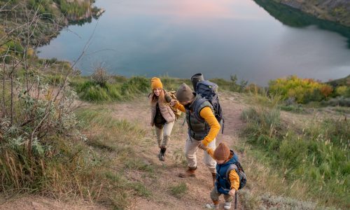 Family of hikers with backpacks climbing mountain against river while bearded man helping his wife and son during trip on weekend